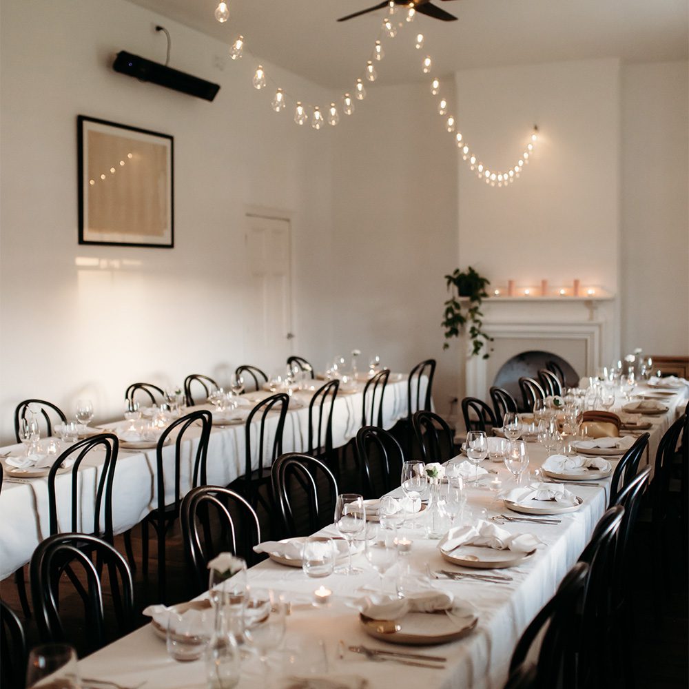 functions-table-1000x1000 A warmly lit dining room set up elegantly for an event, with two long tables covered in white linens, neatly arranged place settings, candles, and string lights hanging from the ceiling. Black curved chairs line the tables, and there's a small fireplace with candles in the background, creating a cosy, intimate atmosphere.